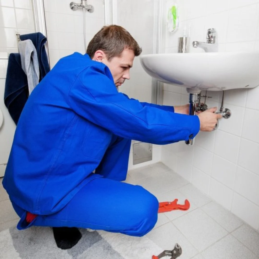 Loodgieter in blauwe overall knielt bij een wastafel en werkt aan de aansluiting