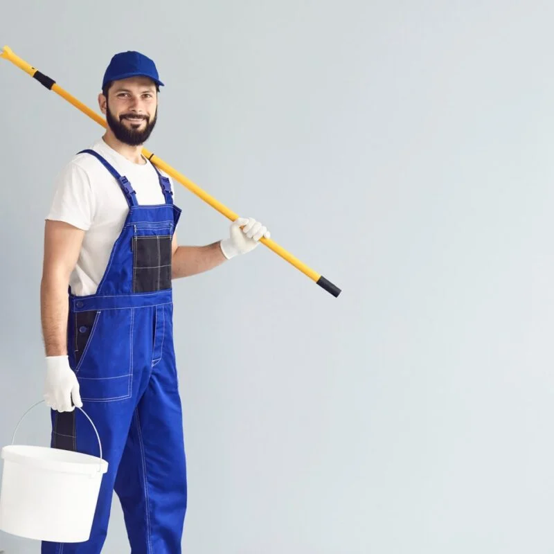 Schilder in blauwe overall met een verfroller en een emmer verf naast een trap